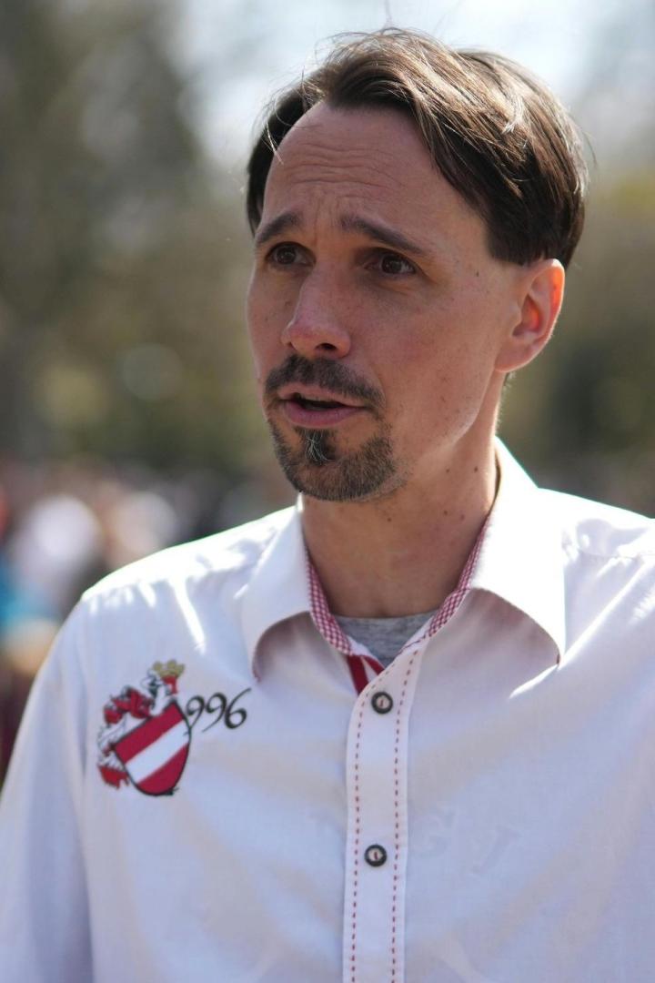 Ein Mann mit Bart gestikuliert in einem weißen Hemd mit Wappen.
