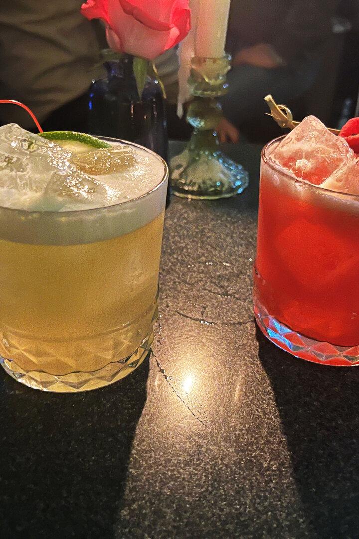 Zwei Cocktails, einer gelb mit Kirsche, der andere rot mit Himbeeren, stehen auf einem Tisch.