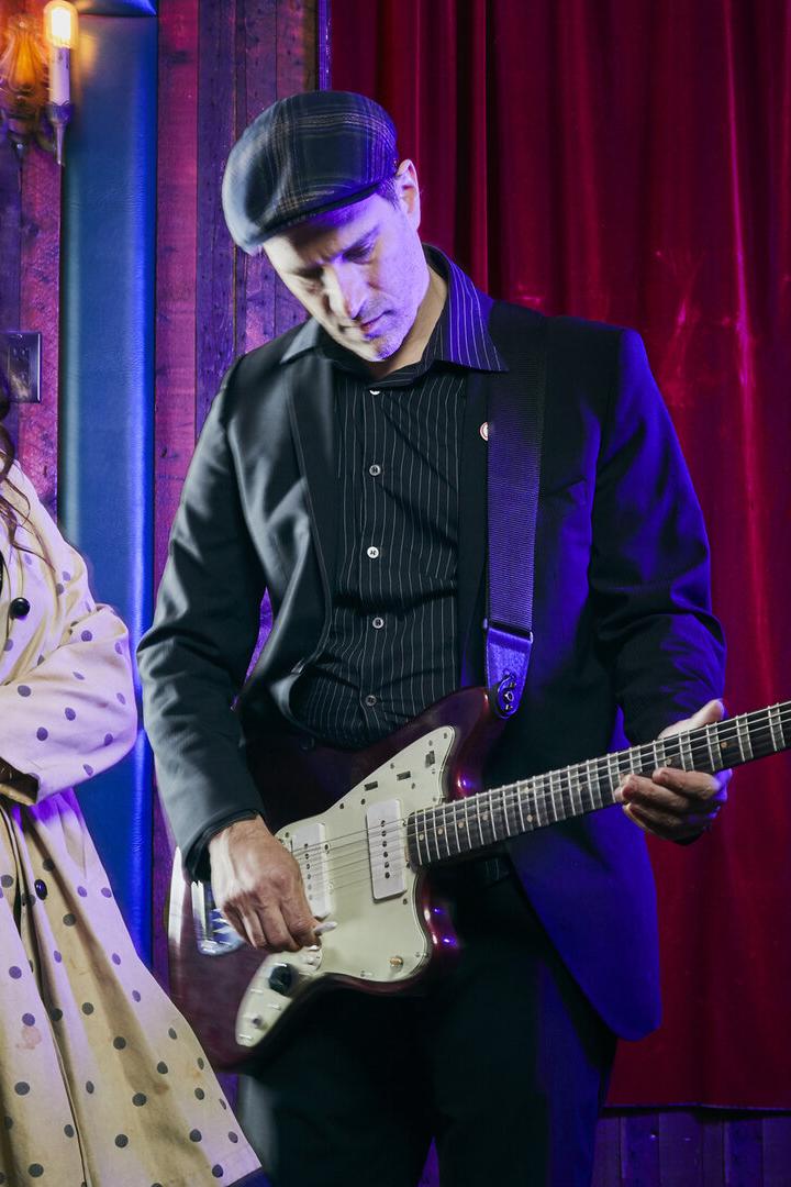 Eine Frau im gepunkteten Mantel posiert neben einem Mann, der Gitarre spielt.