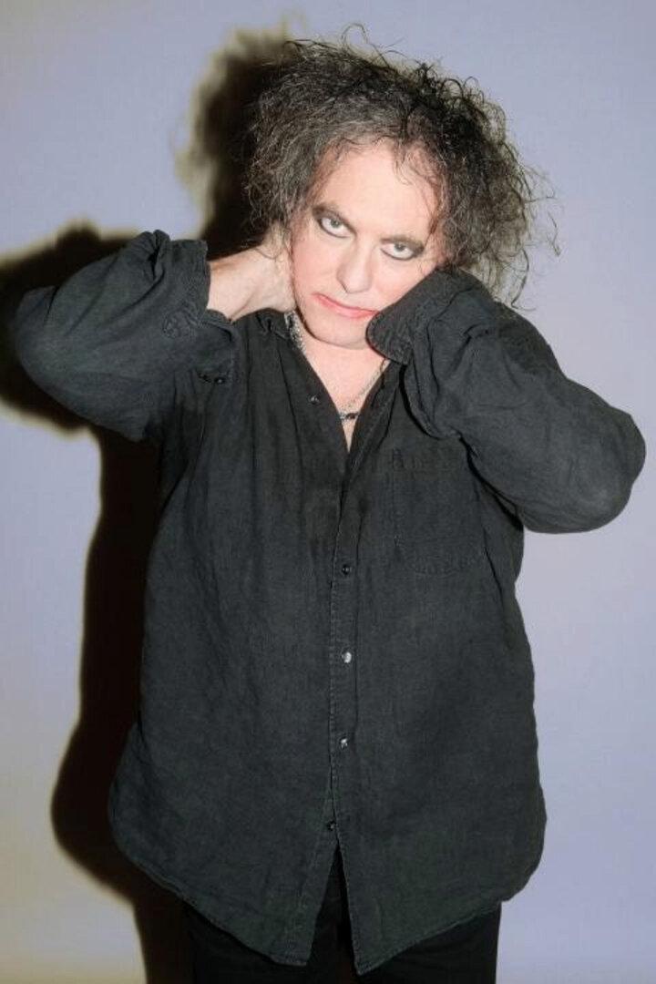 Robert Smith, Sänger der Band The Cure, posiert vor einer grauen Wand.