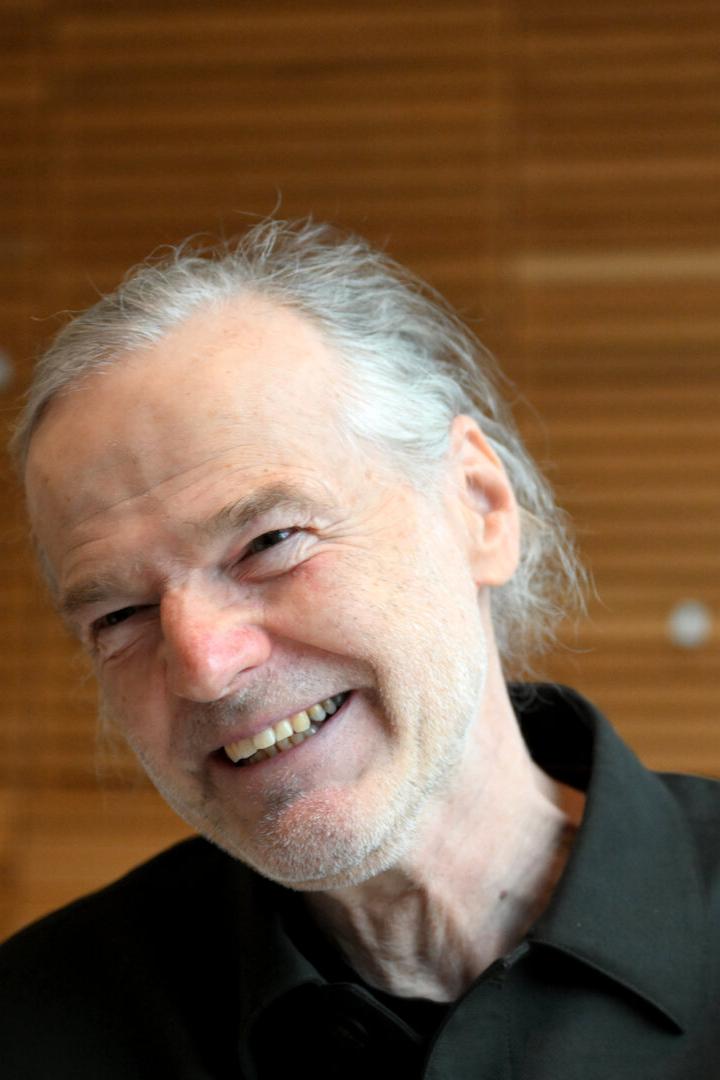 Ein lächelnder älterer Mann mit grauem Haar und schwarzem Hemd vor einer Holzwand.