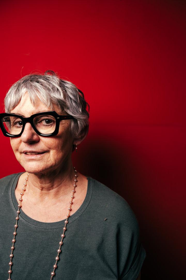 Eine ältere Frau mit grauen Haaren, Brille und einer Perlenkette vor einem roten Hintergrund.