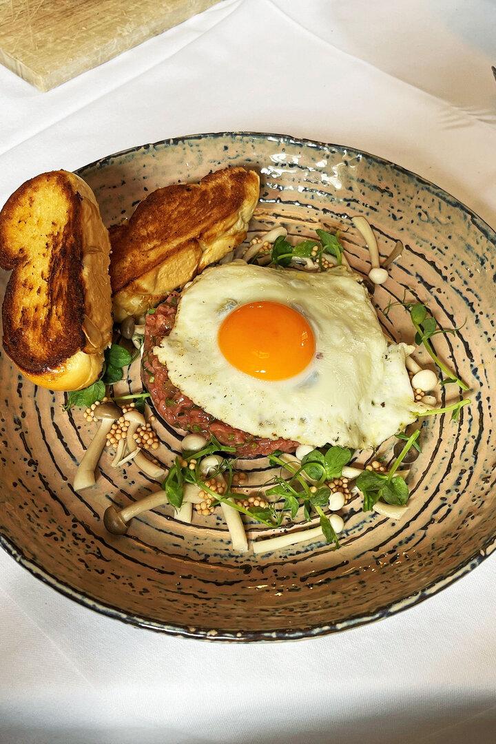 Ein Teller mit Tatar, Spiegelei, Pilzen und geröstetem Brot auf einem Tisch.