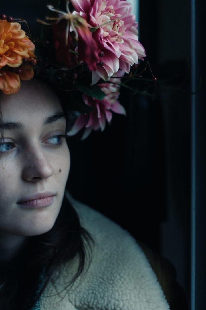 Eine junge Frau mit einem Blumenkranz blickt aus dem Fenster.