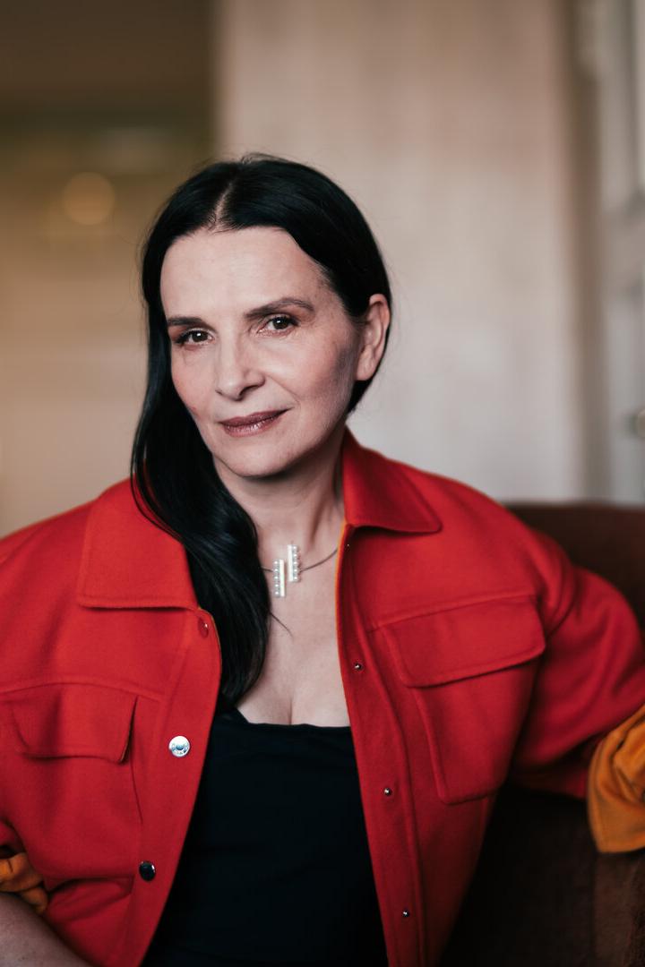 Dunkelhaarige Juliette Binoche in roter Jacke blickt uns direkt an