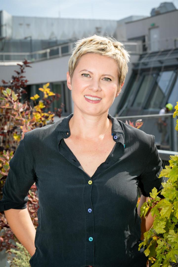 Person mit kurzen blonden Haaren trägt ein schwarzes Hemd mit bunten Knöpfen, steht vor einer Dachterrasse mit Pflanzen