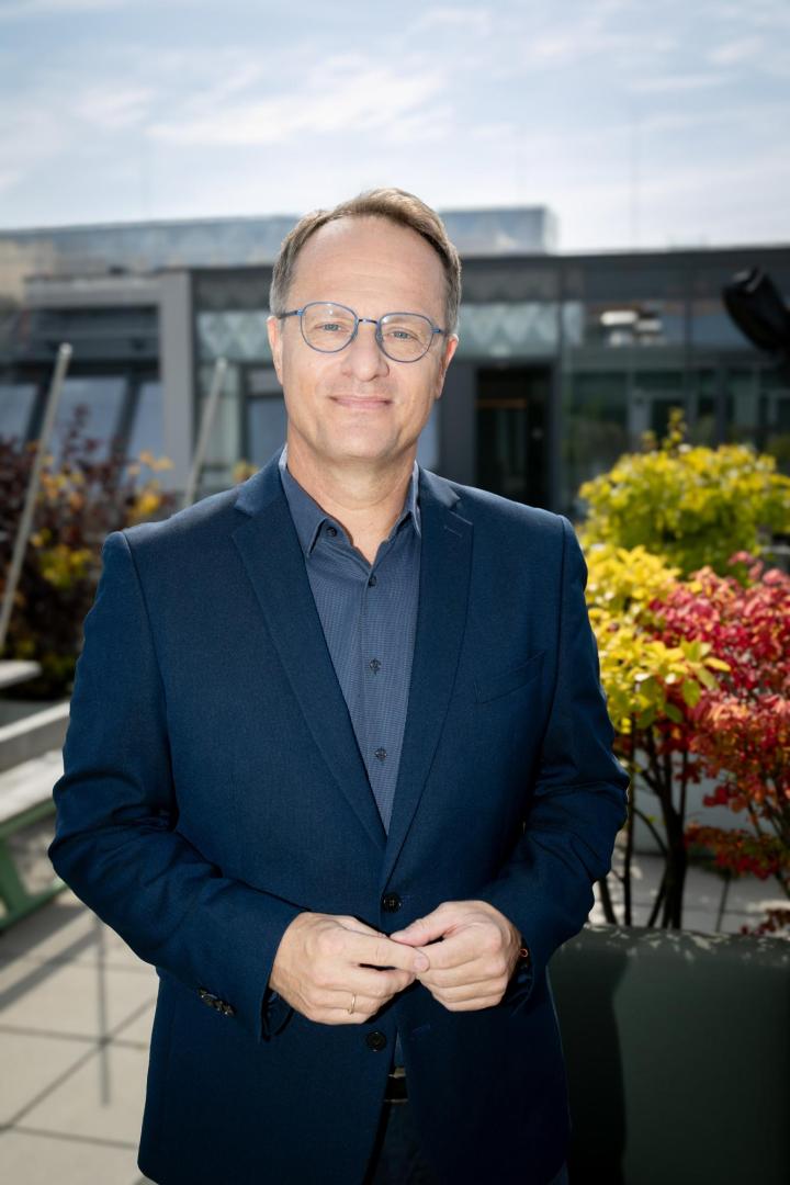 Mann in dunkelblauem Sakko und blauem Hemd steht vor einer Terrasse mit Pflanzen und moderner Glasfassade im Hintergrund.