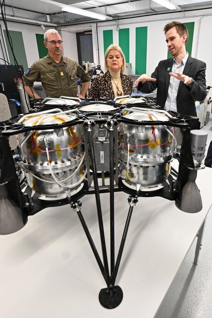 Friedrich Teichmann (li.) und Verteidigungsministerin Claudia Tanner (ÖVP) (Mitte) begutachten die BEACONSAT-Modelle von Moritz Novak (GATE Space) (re.). Im Zentrum des Fotos ist ein metallenes Gerät, das Satellitenmodell zu sehen. Novak deutet mit den Händen, um das Modell zu erklären.