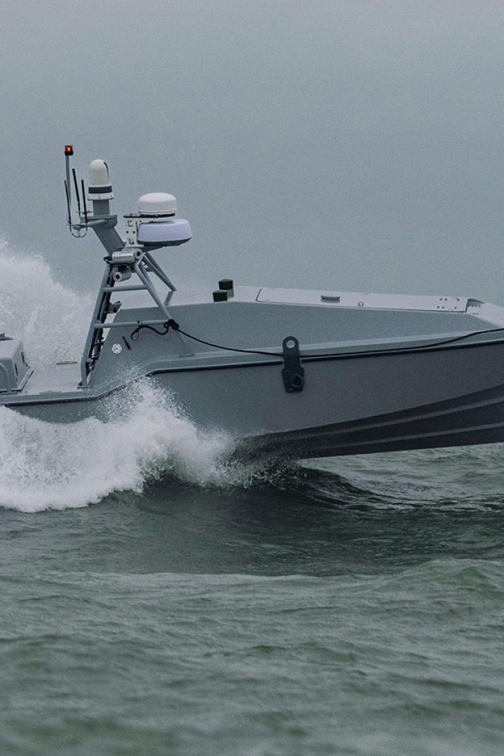 Eine "Corsair"-Drohne (ein graues Motorboot, mit auffälligem Radarsystem) fährt auf dem Wasser und hinterlässt eine große Welle hinter sich.