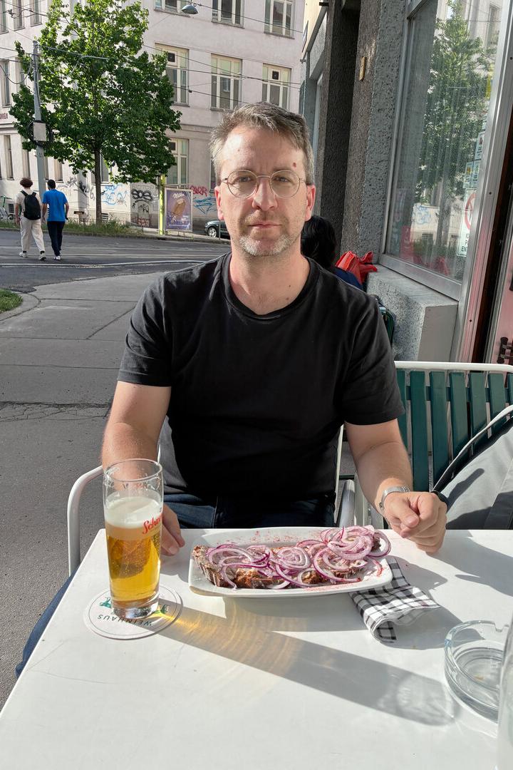 Ein Mann mit Brille und schwarzem T-Shirt sitzt vor einem Schmalzbrot und einem großen Bier