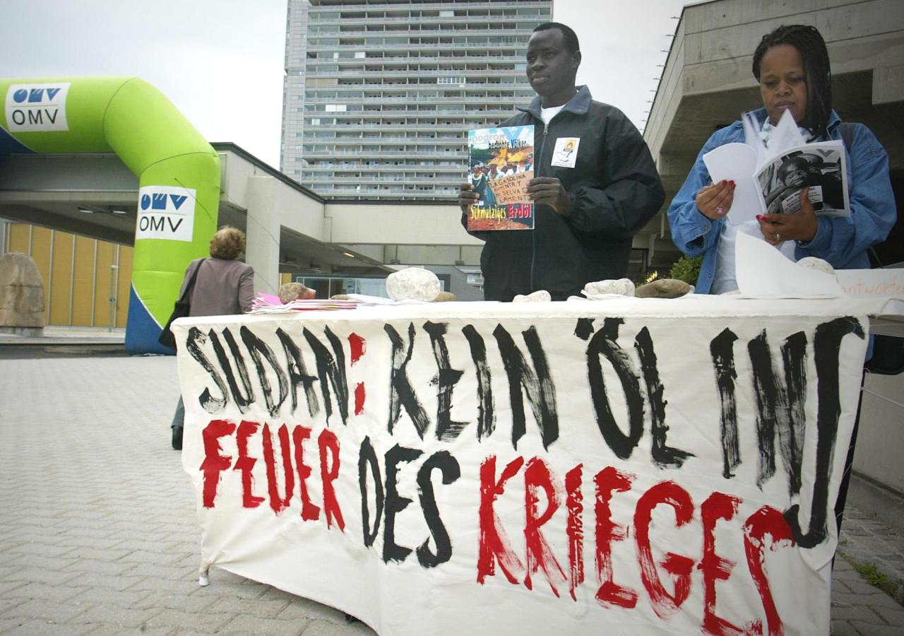 Zwei Aktivisten stehen an einem Stand mit der Aufschrift „Sudan: Kein Öl ins Feuer des Krieges“.