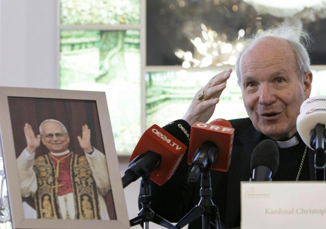 Kardinal Christoph Schönborn spricht vor Mikrofonen neben einem Bild von Papst Johannes Paul II..