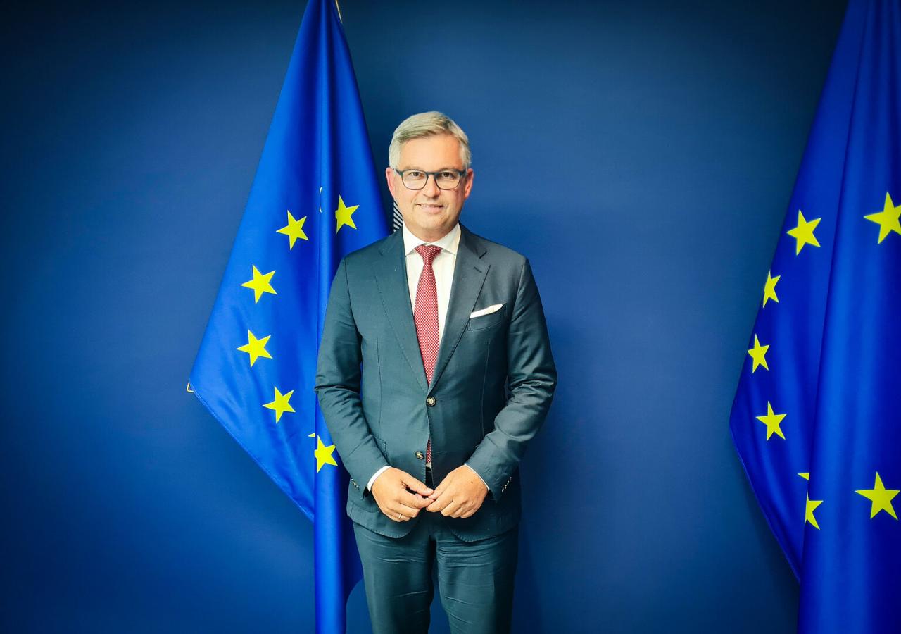 Migrationskommissar Magnus Brunner (ÖVP) steht vor einem blauen Hintergrund und zwei EU-Flaggen. Er ist am Rande der Plenarsitzung des Europäischen Parlaments Anfang September 2025 in Straßbourg.