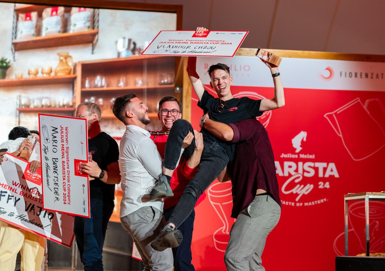 Eine Gruppe von Männern feiern in der Eventalle von Julius Meinl. Einer wird  hochgehoben. Alle halten große Schilder mit Texten. Im Hintergrund eine Küchenzeile.