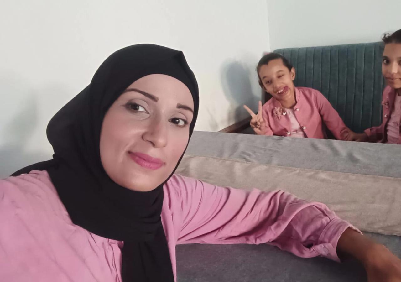 46-219477863 Eine Frau in Kopftuch posiert vor einem Esstisch für ein Selfie, am anderen Ende des Tische sitzen zwei Mädchen.