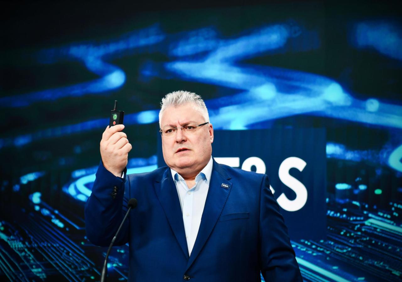 Michael Mertin hält eine Fernbedienung mit seiner rechten Hand hoch. Hinter ihm ist eine Vidi-Wall mit dunkelblauen und hellblauen Kabeln und Elektronik darauf.