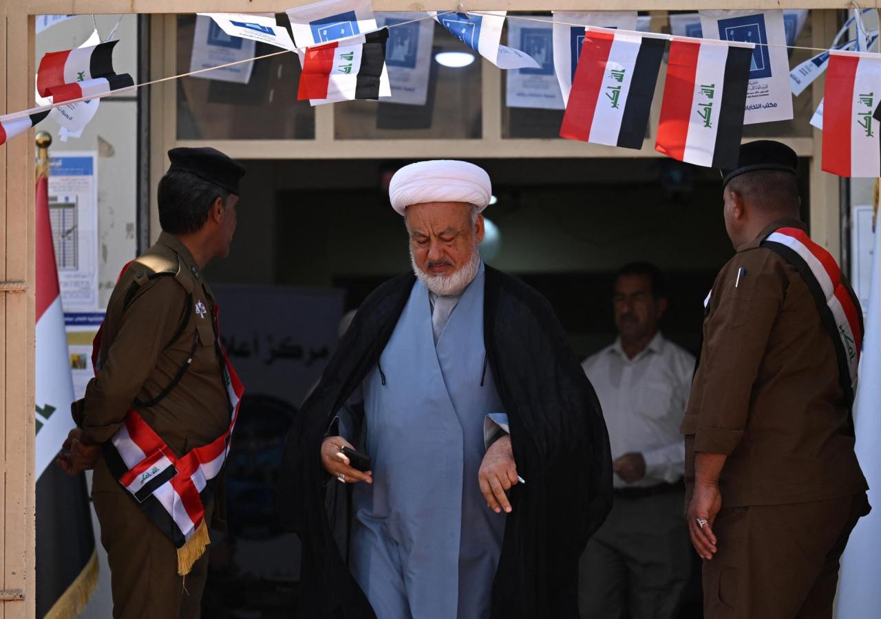 Ein Mann in Turban verlässt ein mit Irak-Flaggen drapiertes Wahllokal, daneben stehen Sicherheitsoffiziere.