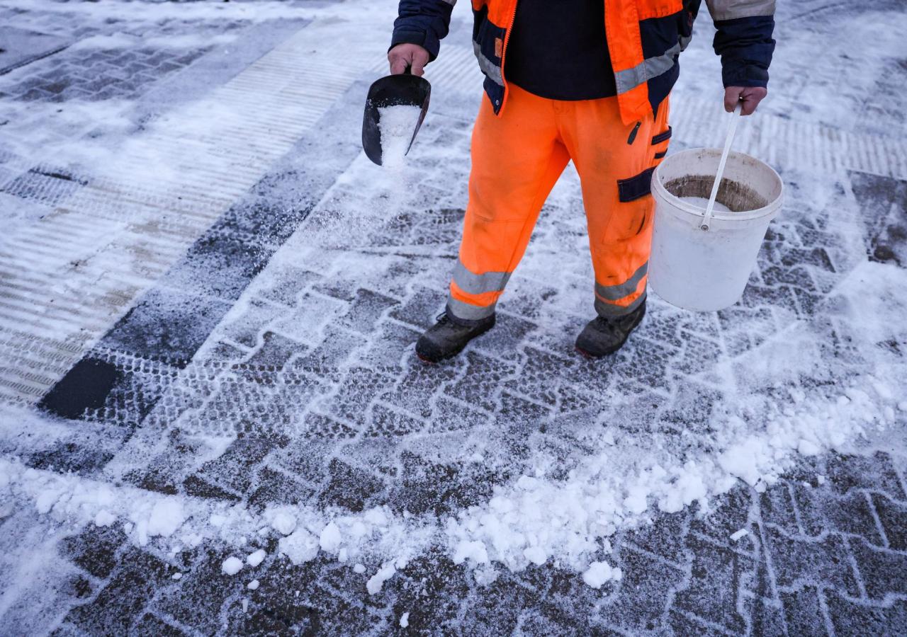 Ein Mitarbeiter des Winterdienstes verteilt Streusalz bei Regen und Schneeglätte auf den Fußweg an einer Straßenkreuzung. 