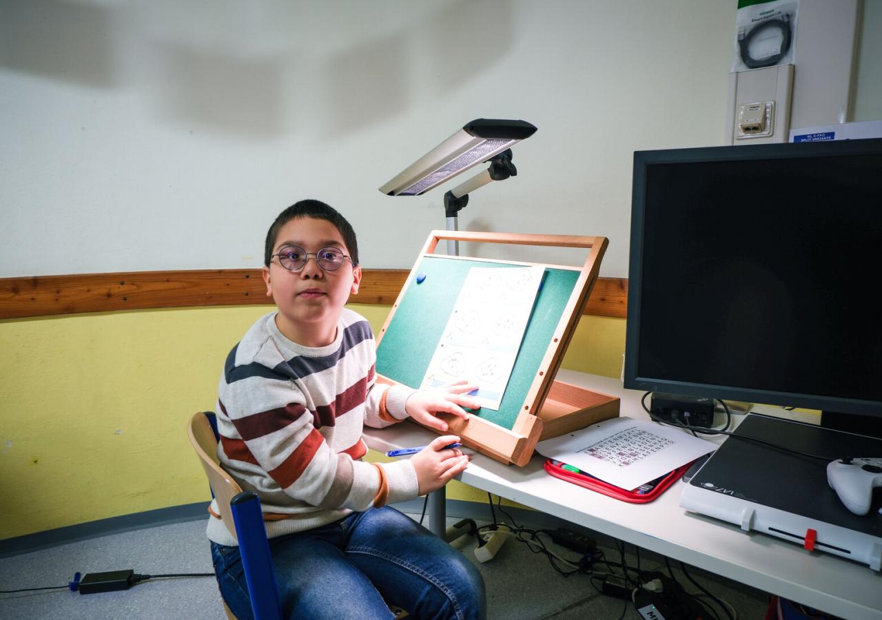 Ein Kind sitzt an einem Schreibtisch in einem Klassenraum und arbeitet an einer geneigten Lern- oder Lesetafel. Auf dem Tisch stehen weitere Lernmaterialien und ein großer Monitor.