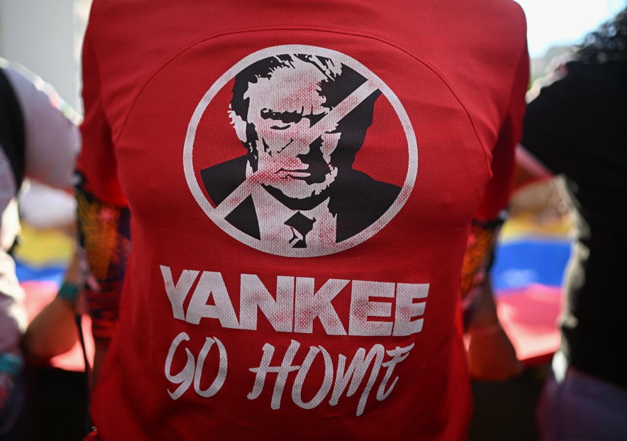 Eine Person trägt ein rotes Shirt mit der Aufschrift "Yankee Go Home" und dem durchgestrichenen Konterfei von Donald Trump, dahinter eine Venezuela-Flagge