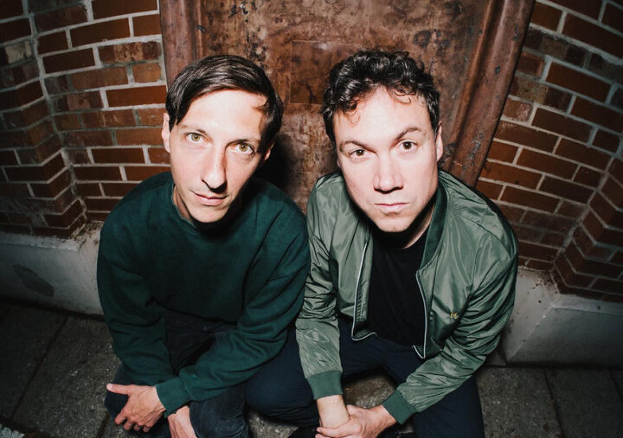 Zwei junge Männer in einer Art Keller blicken mit ernsten Gesichtern in die Fischaugen-Kamera 