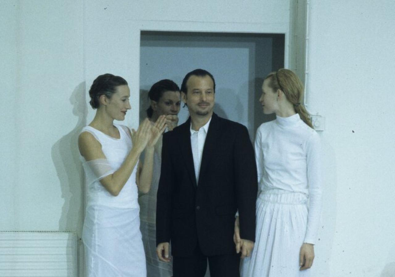 Mann im dunklen Anzug, umstellt von Frauen in weißen Kleidern