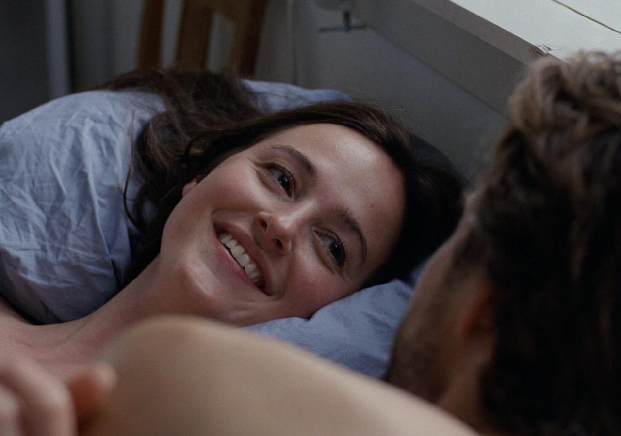 Lächelnde junge Frau liegt in einem Bett, blickt einen Mann an, den man nur von hinten sieht. 