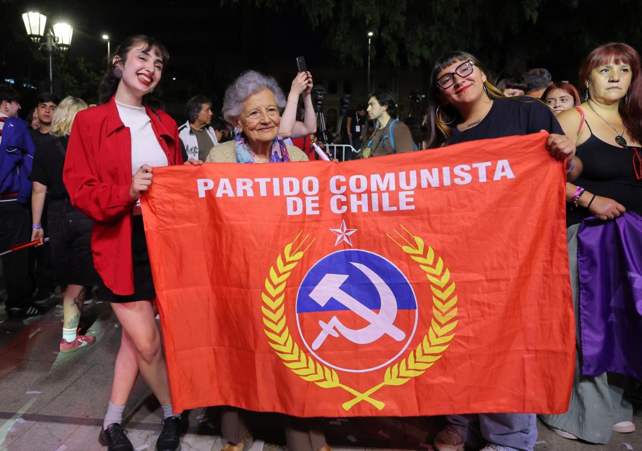 Drei Personen (zwei Junge und eine ältere Frauen) halten eine rote Flagge mit der Aufschrift "partido communista de chile" mit Hammer und Sichel hoch.