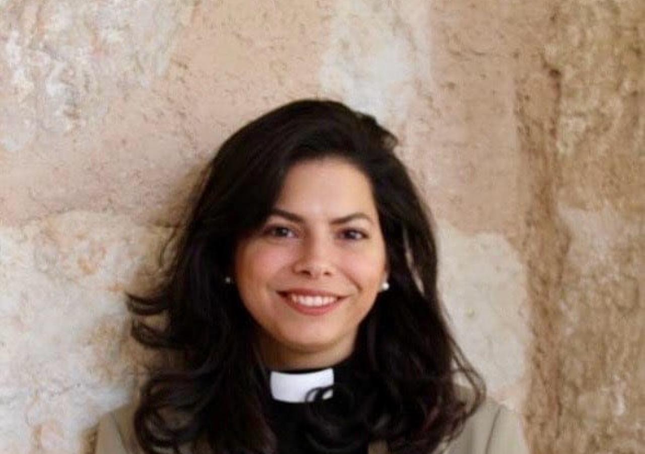 Sally Azar steht in Priestergewand vor einer Steinwand.