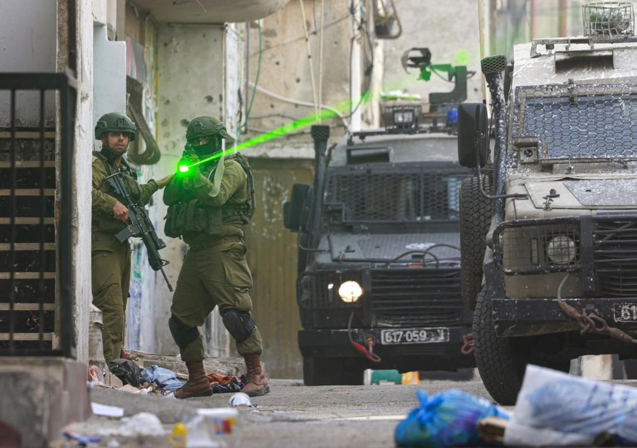 Zwei israelische Soldaten, einer zielt mit seinem Gewehr, ein Laser ist sichtbar, rechts zwei gepanzerte Autos.