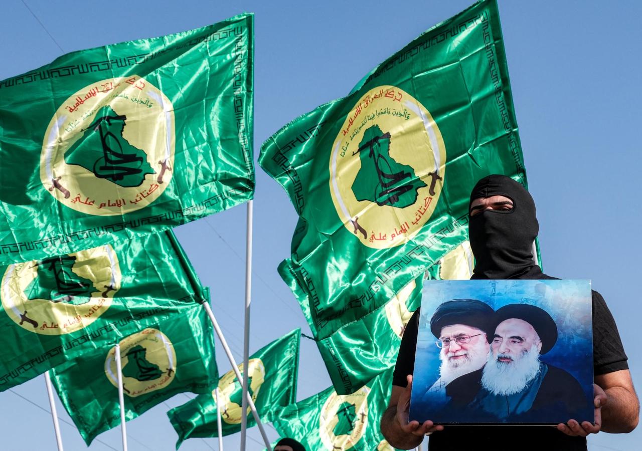 Ein Mann hält ein Bild mit Khomeini und einem irakischen Geistlichen hoch, dahinter grün-gelbe Flaggen einer proiranischen Miliz.