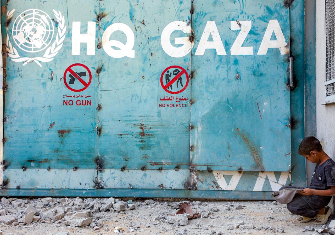 Ein Junge kniet vor einem eisernen Tor mit der Aufschrift "HQ GAZA" und dem UN-Logo.