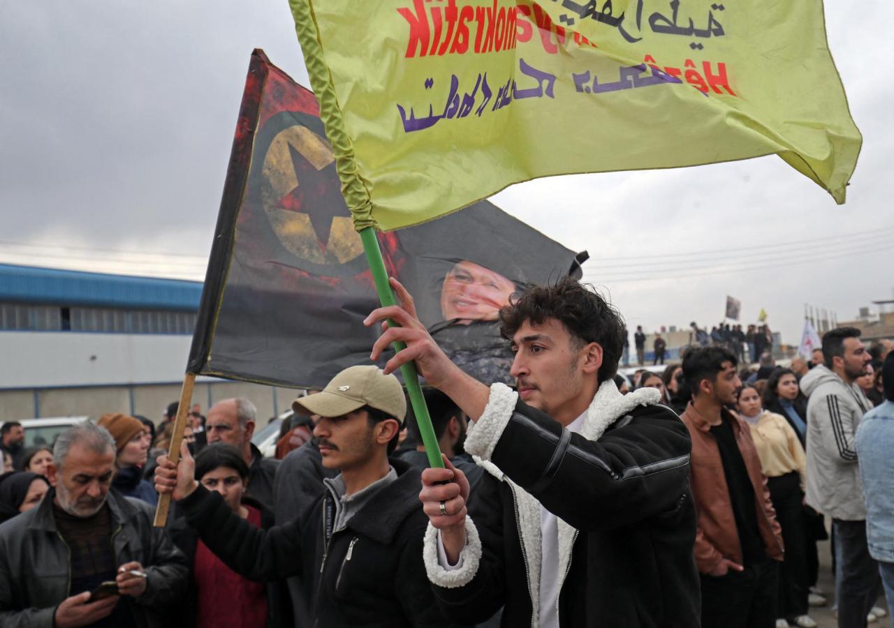 Ein Mann schwenkt eine Fahne der SDF, dahinter eine Flagge mit PKK-Logo.