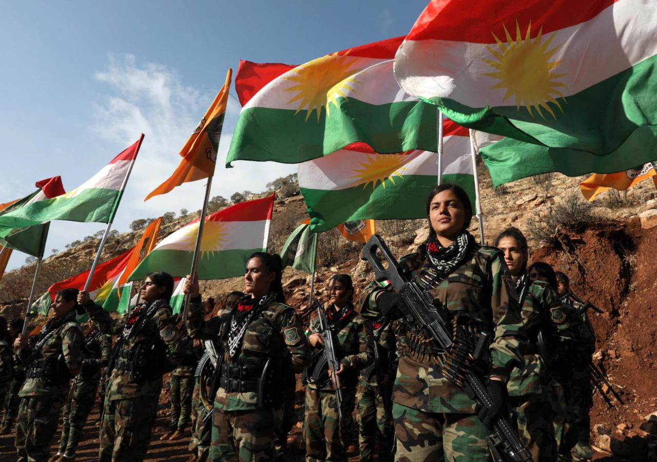 Frauen in Tarnkleidung stehen mit Gewehren vor kurdischen Flaggen.