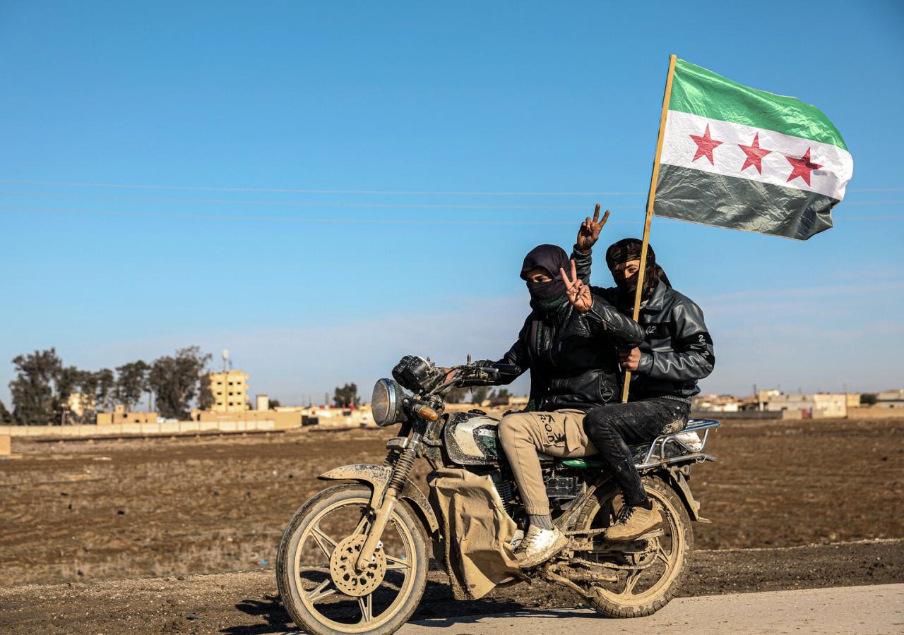 Zwei Männer auf einem Motorrad zeigen Peace-Zeichen, einer hält eine syrische Flagge.
