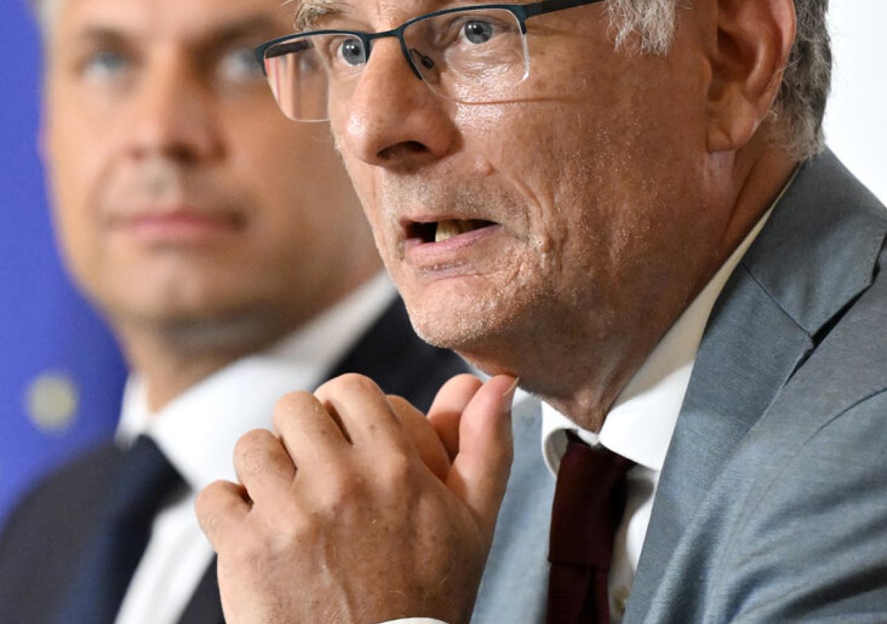Ein Mann mit Brille spricht, während ein anderer im Hintergrund steht.