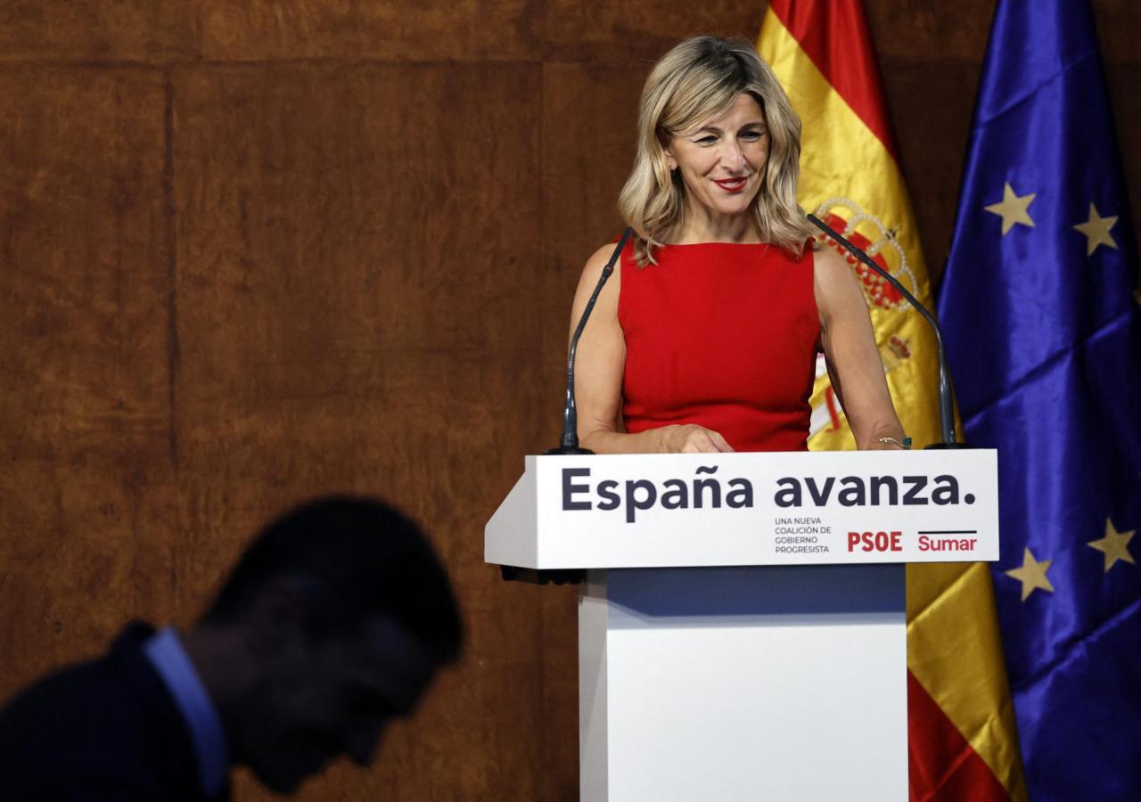 Yolanda Díaz vor einem Rednerpult mit der Aufschrift (Espana avanza.), links unten sieht man einen in Schatten gehüllten Mann.