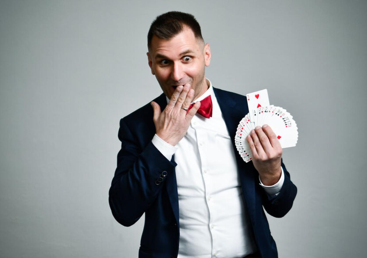 Ein Mann im Anzug mit roter Fliege hält lächelnd ein Fächer aus Spielkarten in der Hand.
