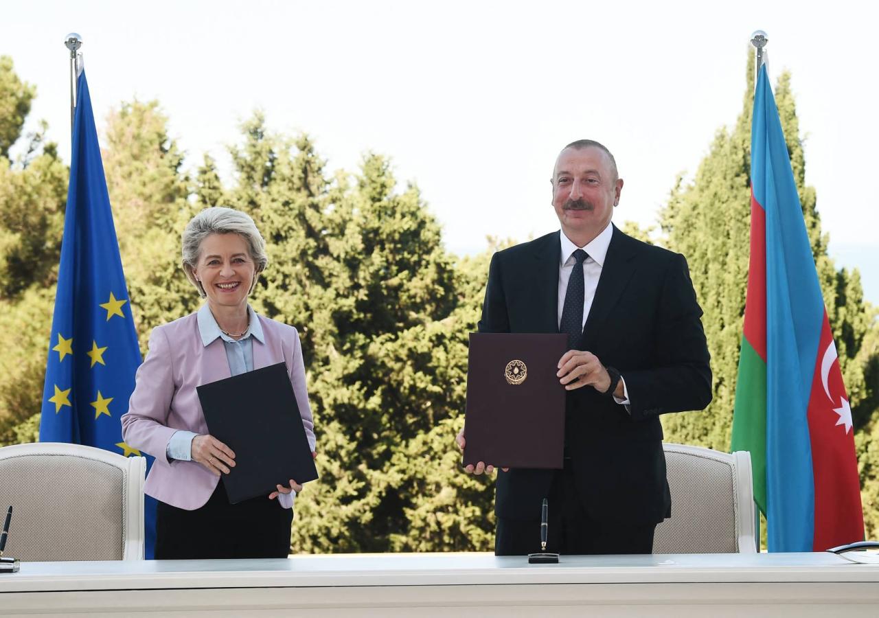 Ursula von der Leyen und Ilham Aliyev halten vor Bäumen zwei Mappen hoch. Sie lächeln beide.