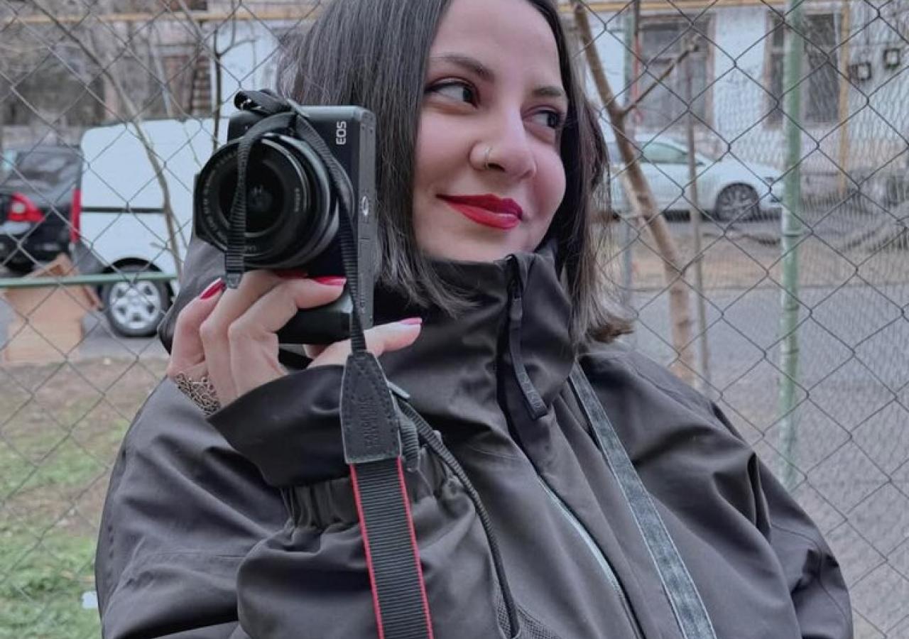 Guliyeva posiert mit einer Kamera vor einem Maschendrahtzaun.