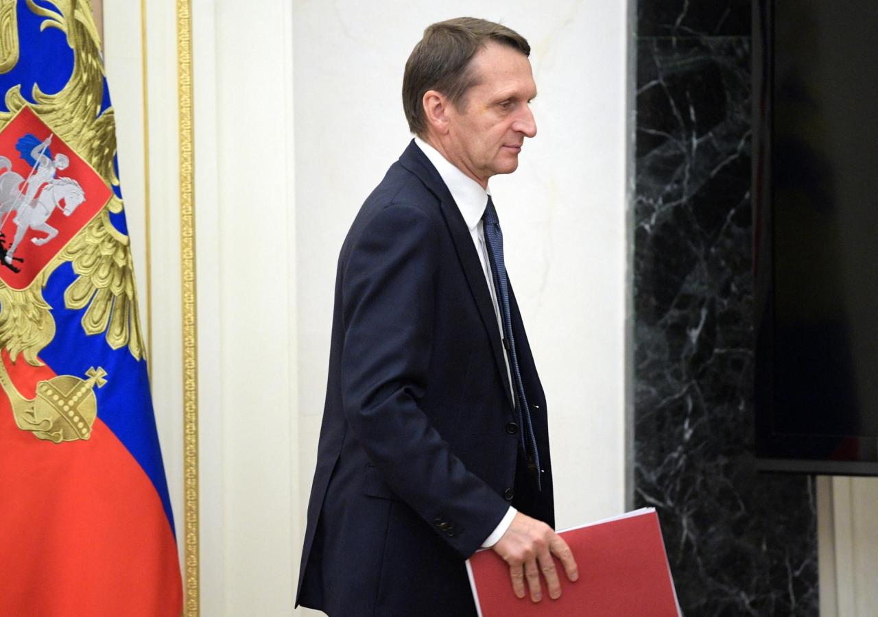 Sergei Jewgenjewitsch Naryschkin vor russischer Flagge, er geht nach rechts mit einer roten Mappe in der Hand.