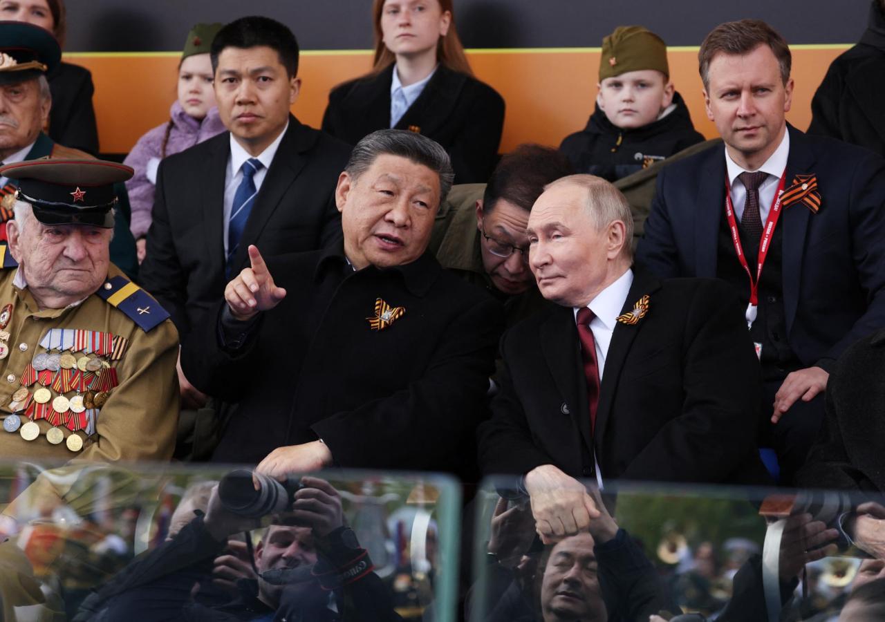 Xi Jingping und Wladimir Putin sitzen auf einer Tribüne, Jingping zeigt mit seinem Zeigefinger auf etwas, Putin hört zu.