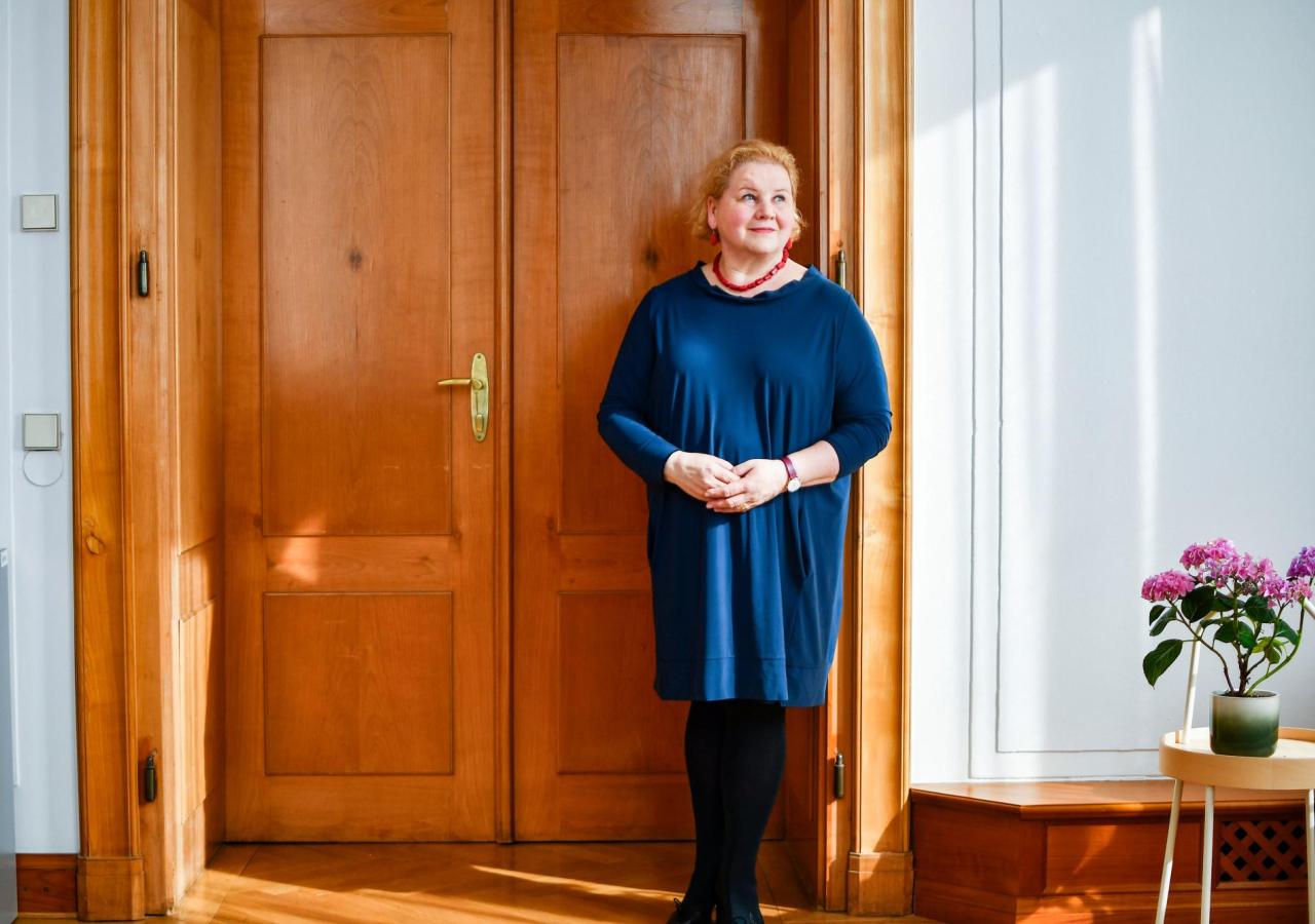 Eine Frau im blauen Kleid, es ist Sozialministerin Schumann, steht vor einer braunen Tür