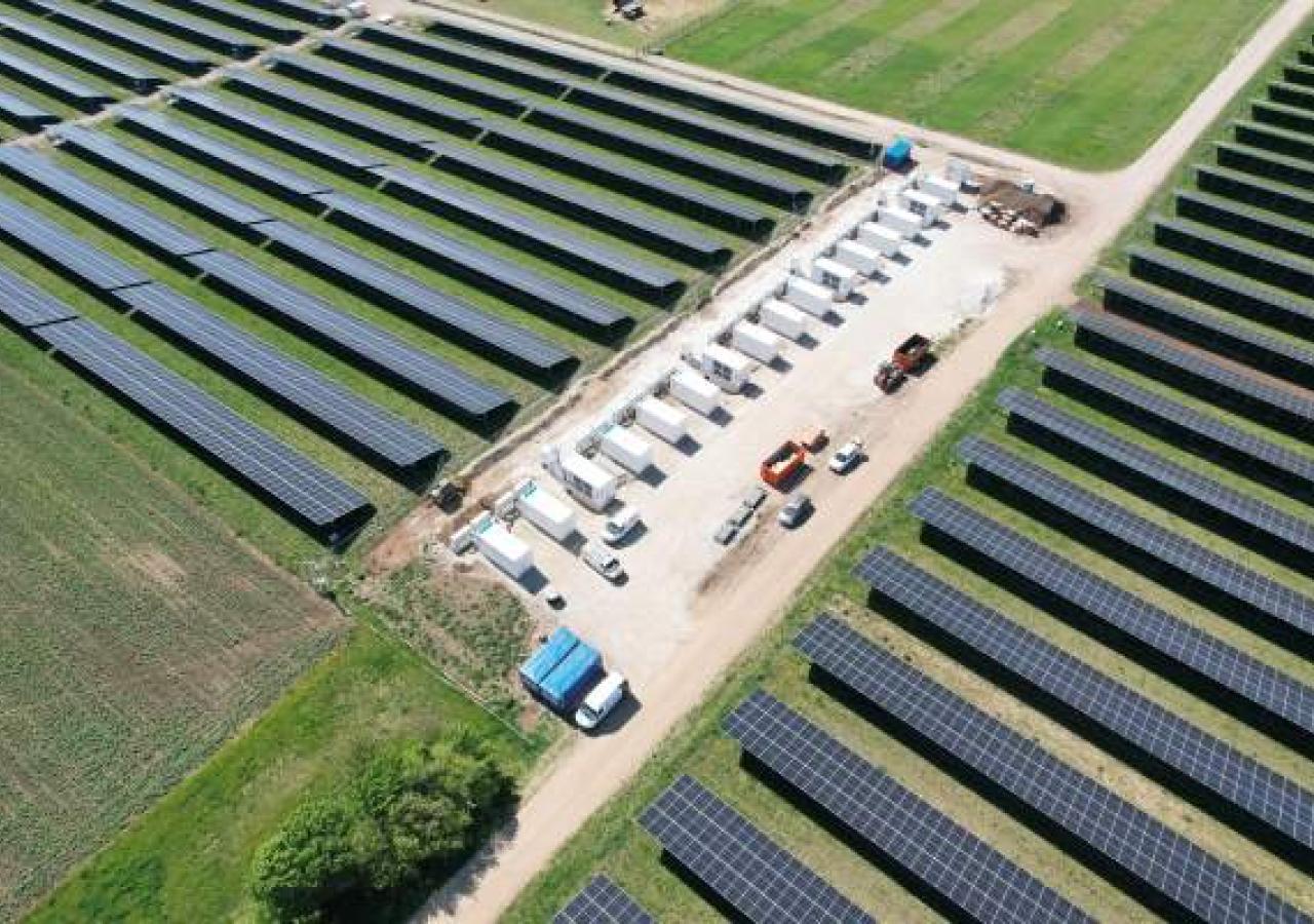 Luftaufnahme einer Freiflächen-Solaranlage mit mehreren Reihen von Solarmodulen und einem zentralen Bereich mit weißen Batteriespeichern und Fahrzeugen