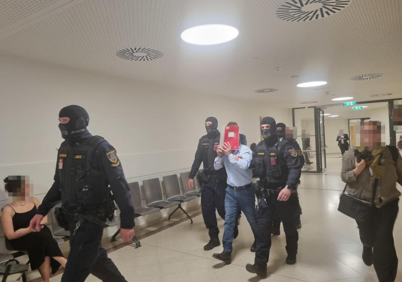 H. verdeckt sein Gesicht mit einem Notizheft, rundherum Polizisten und Journalisten.