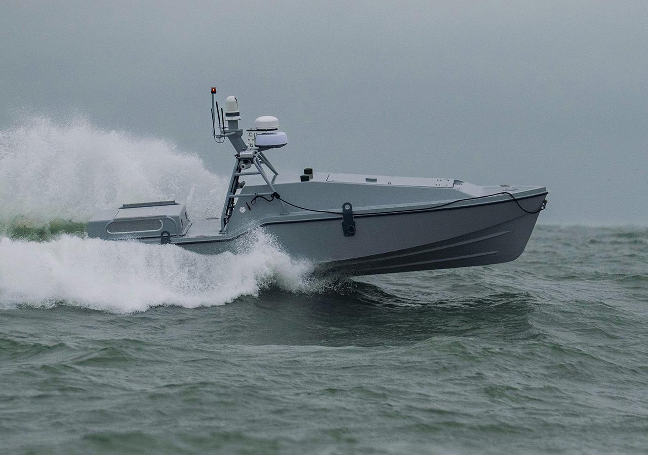 Eine "Corsair"-Drohne (ein graues Motorboot, mit auffälligem Radarsystem) fährt auf dem Wasser und hinterlässt eine große Welle hinter sich.