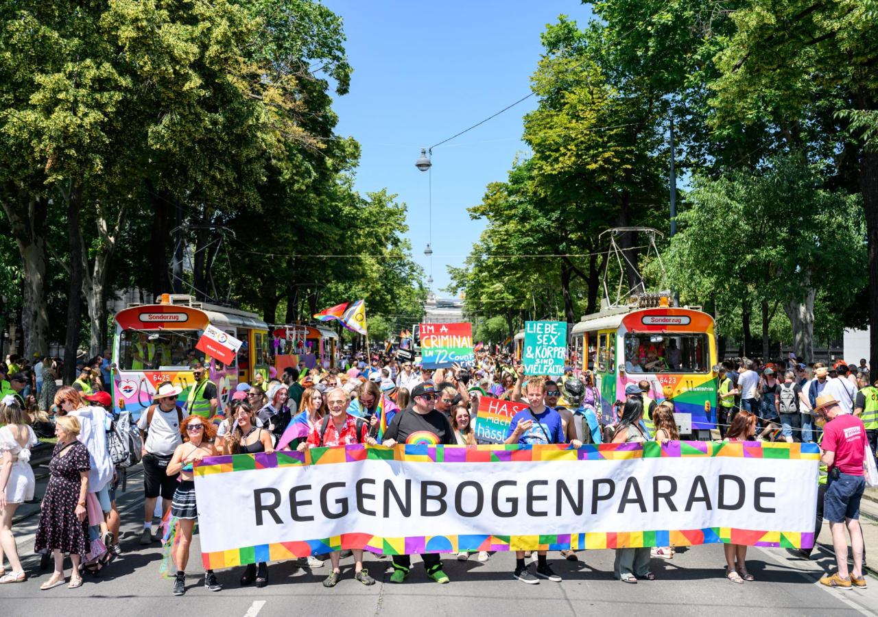Viele Protestierende auf die Vienna ride Regenbogenparade halten einen großen banner. Im Hintergrund sind bunte Fahnen.