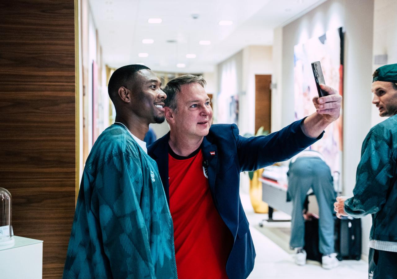 David Alaba lächelt, während ein Mann in einem roten Trikot und blauem Sakko, Sportminister Babler, ein Selfie mit ihm in einem Flur macht.