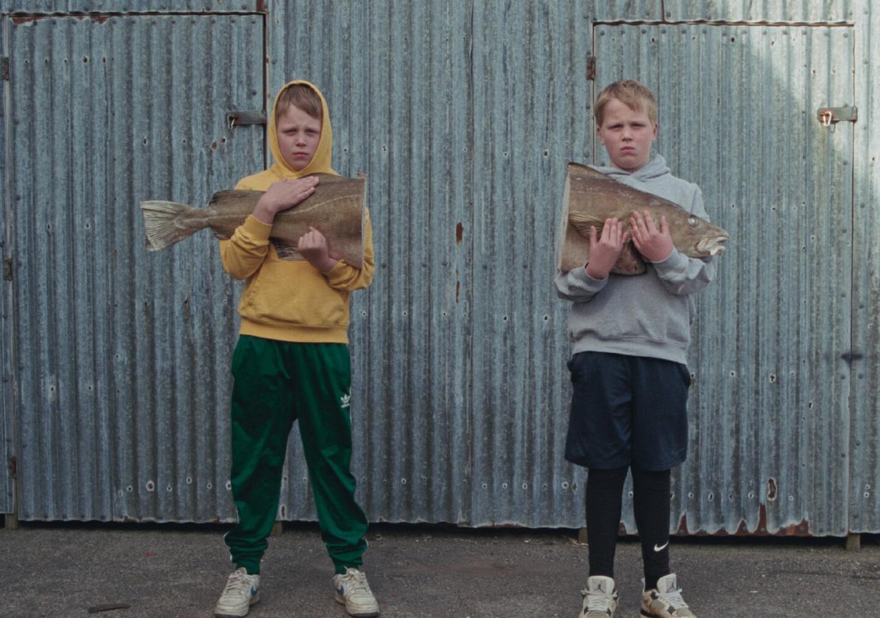 Zwei Buben stehen vor einem Wellblechtor, jeder hält einen großen halbierten Fisch in den Atmen, an die Brust gedrückt