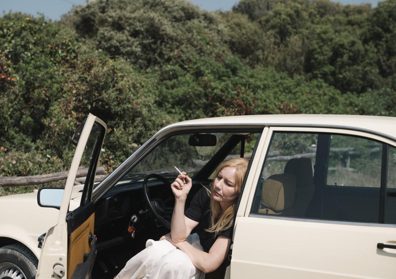 Die Schauspielerin Sandra Hüller mit blonder Perücke, rauchend, in einem geparkten Auto sitzend, im Hintergrund ein Wald
